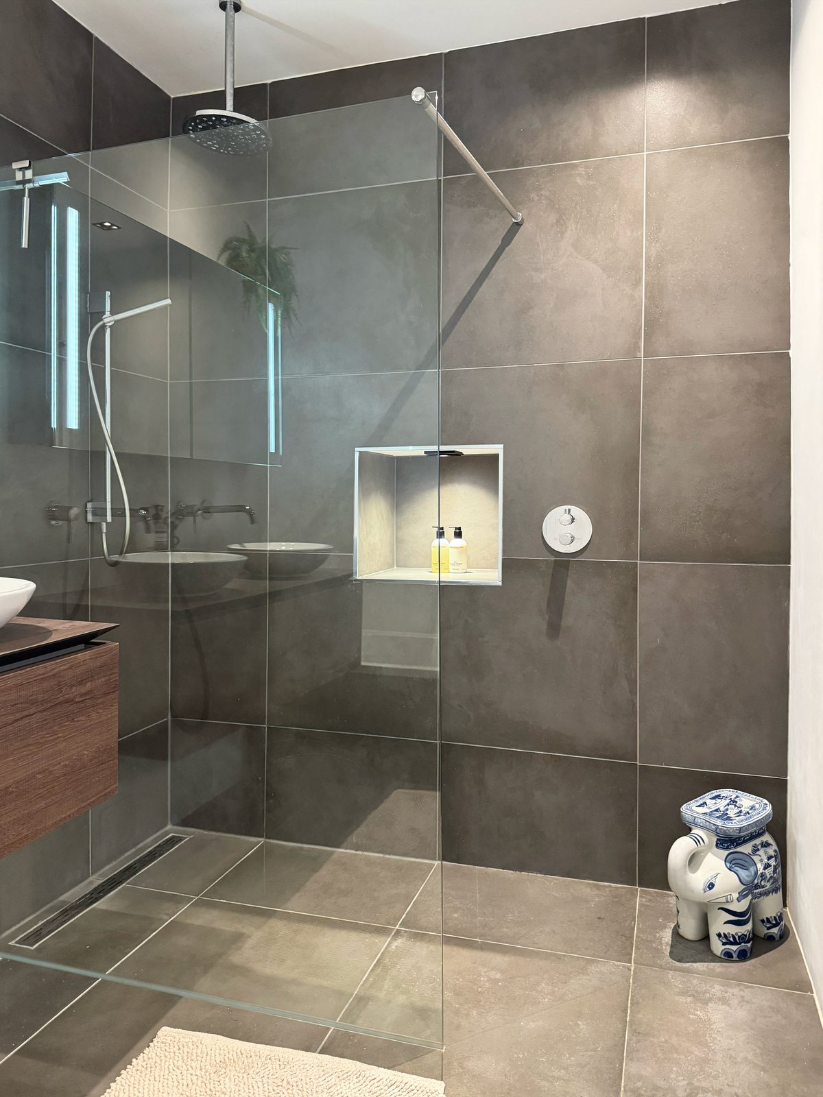 The walk-in rain shower at Waal in large grey stone tile, with a ceiling rain head, a recessed niche and a Delft blue stool