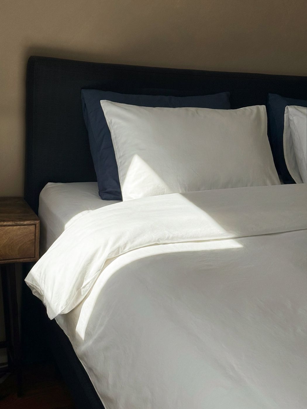 Crisp white percale bed linen at Waal in morning sunlight, layered against navy pillows and a dark headboard