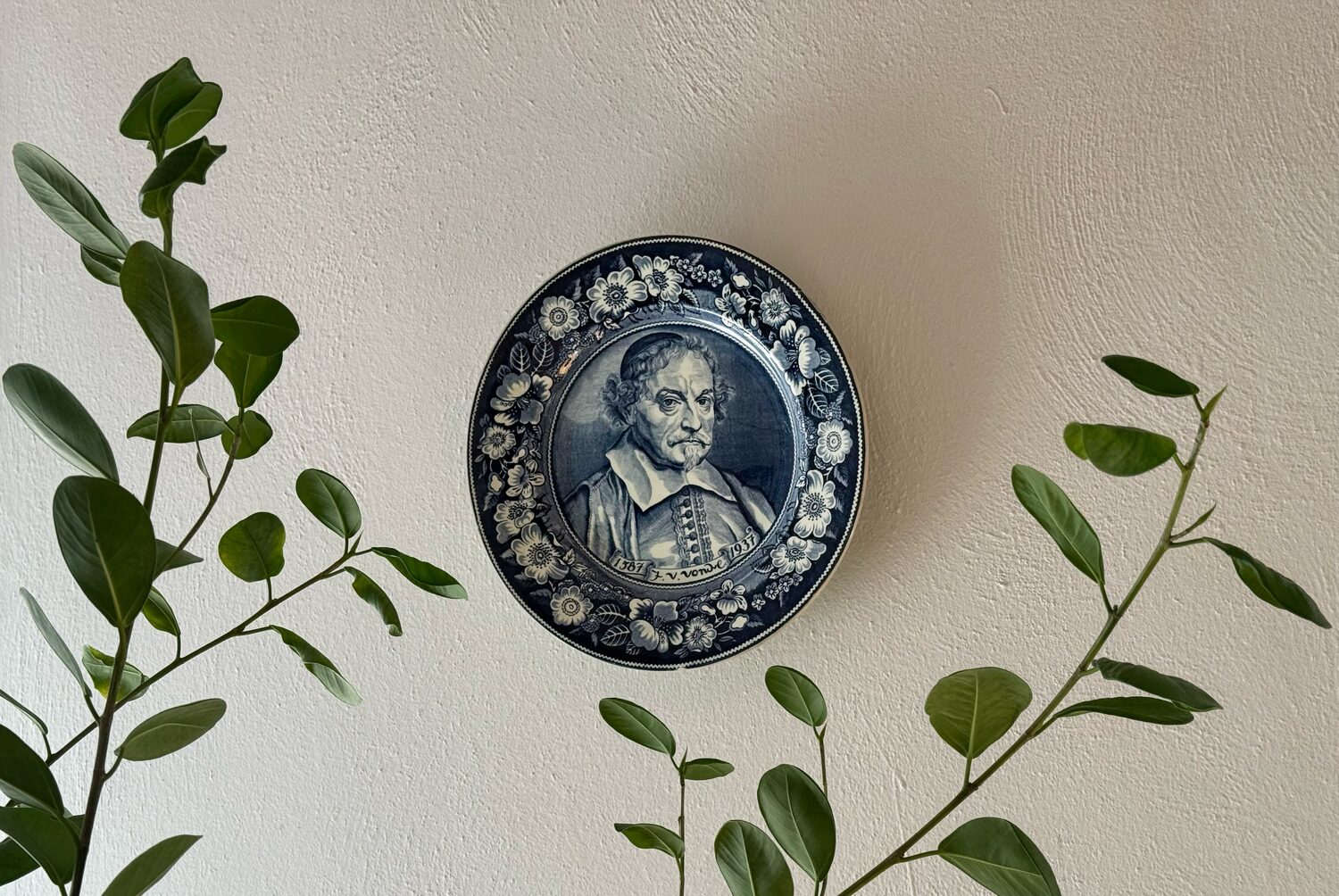 Delft blue commemorative plate hung on a white wall between green branches at Waal