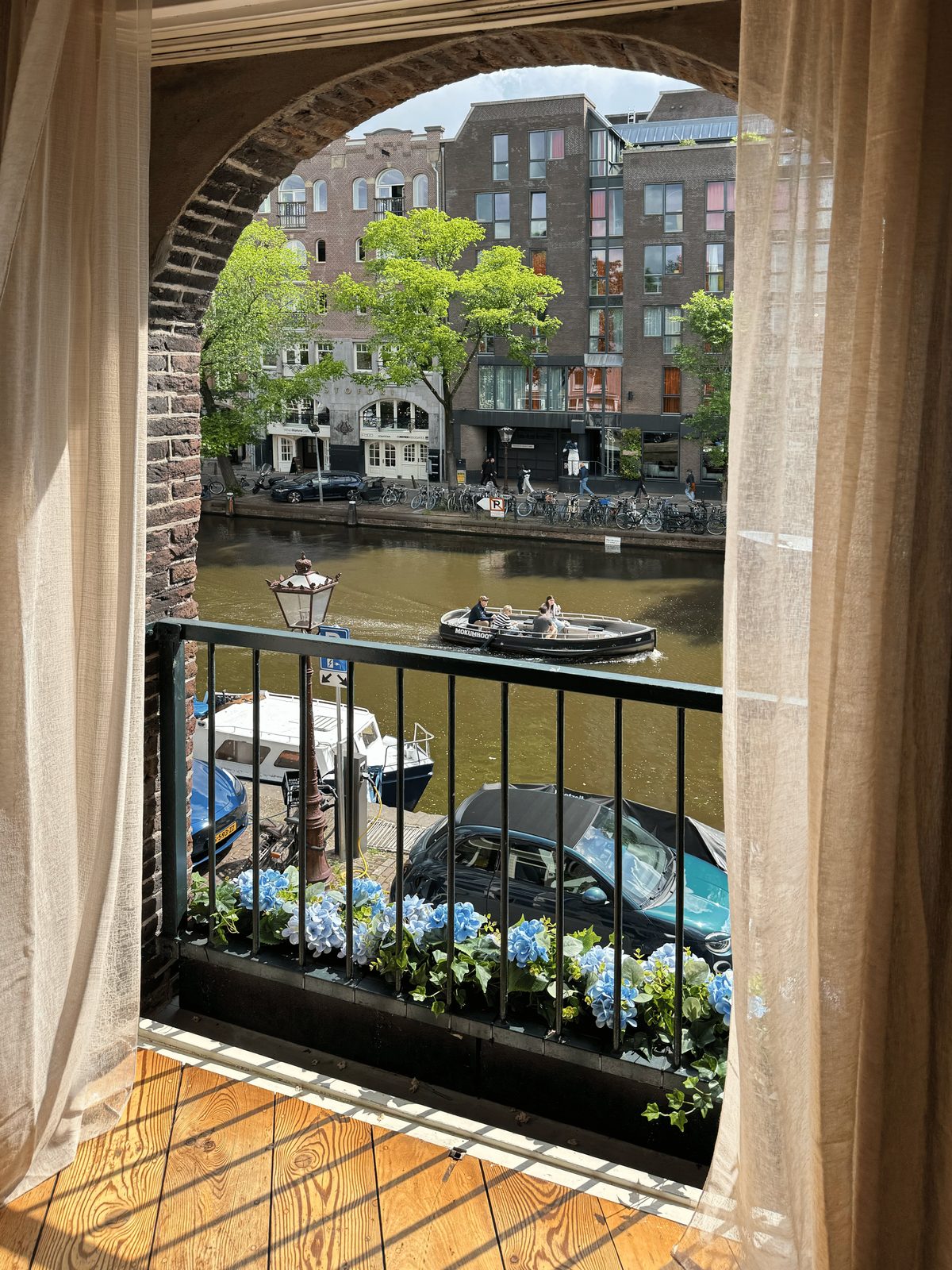 The view from the French balcony at Waal through an arched window, with sheer curtains, a window box of blue hydrangeas and a boat passing on the Prinsengracht