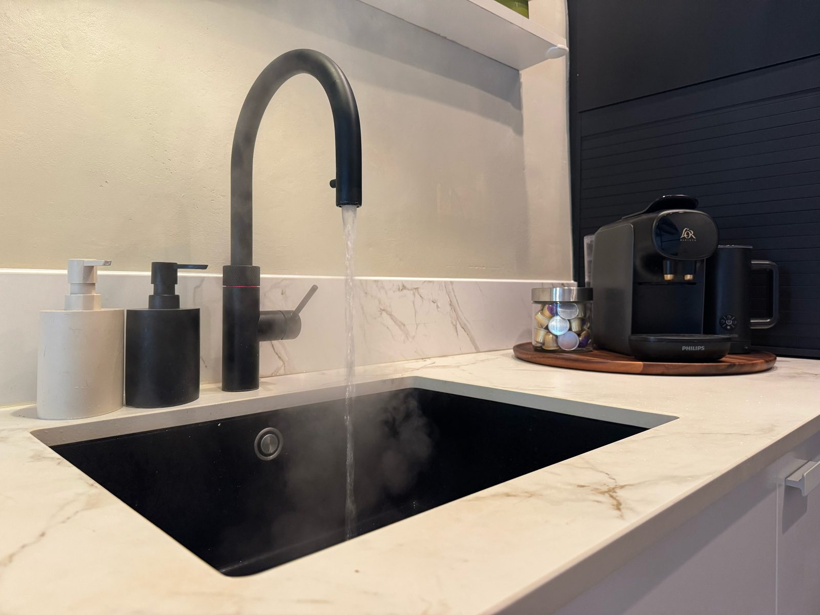 Black Quooker tap running over a matte sink on the marble counter at Waal, beside a coffee machine and capsules