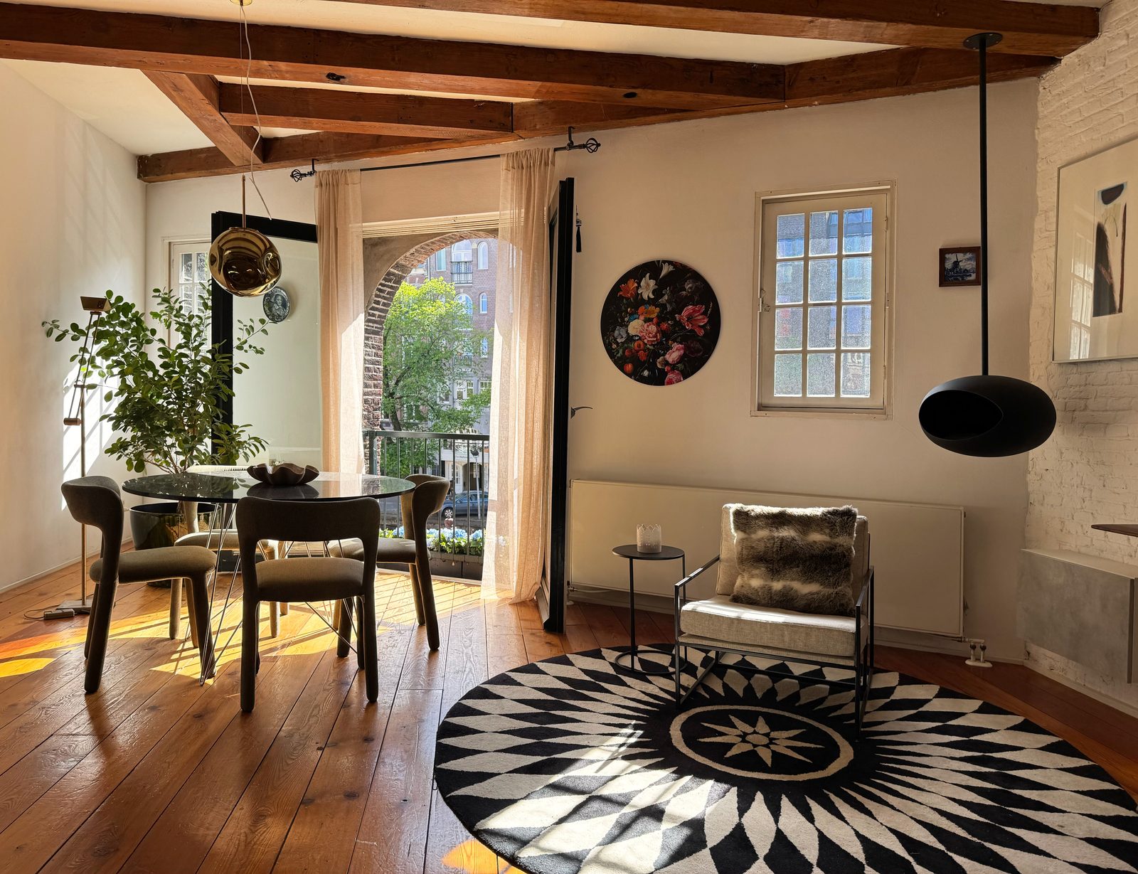 Open living and dining room at Waal under exposed beams, with a round graphic rug, dining table and French balcony