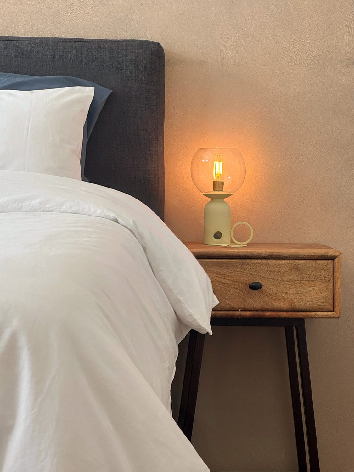 A bedside detail in the master at Waal: a warm glass lamp glowing on a mango-wood nightstand beside crisp white and navy linen