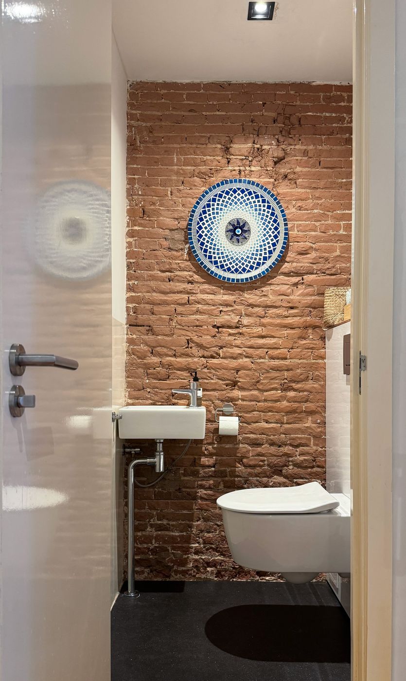 The guest powder room at Waal with an exposed red brick wall, a blue mosaic mirror and a wall-hung basin and toilet
